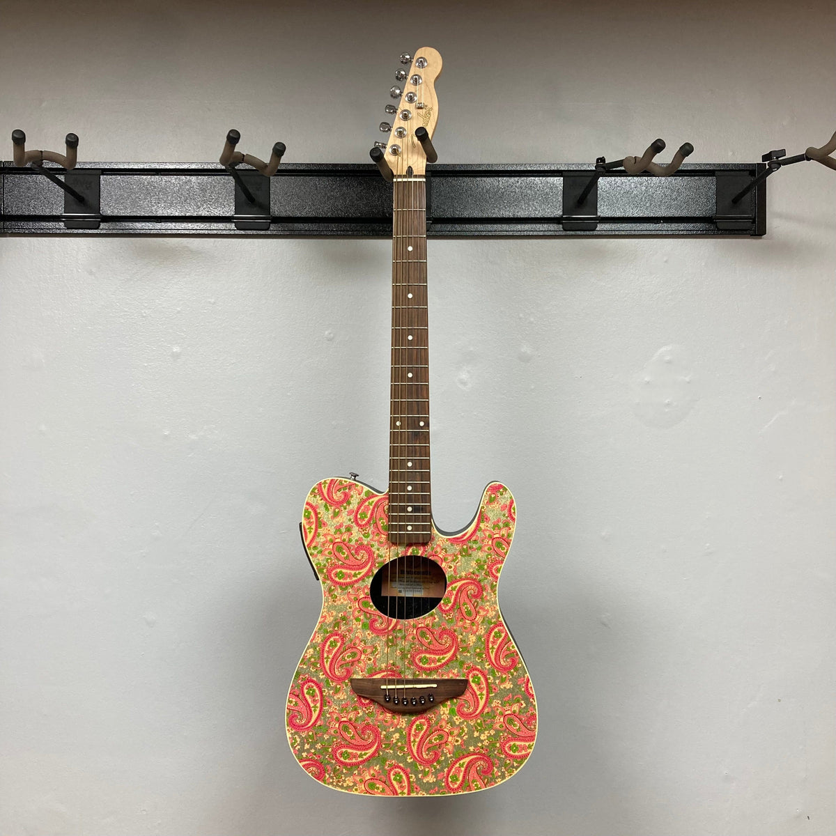 Fender Stratacoustic Pink Paisley guitar with intricate design, hanging on a hook. Features slim body and built-in electronics for versatile playing.