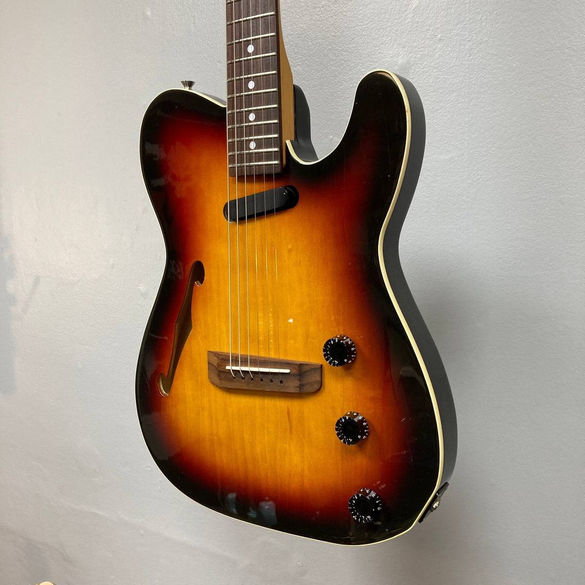 Fender Telecaster TLAC MIJ Acoustic/Electric Hybrid 1993-1997 3 Color Sunburst, hanging on a wall, showcasing its strings and black dials with white numbers.