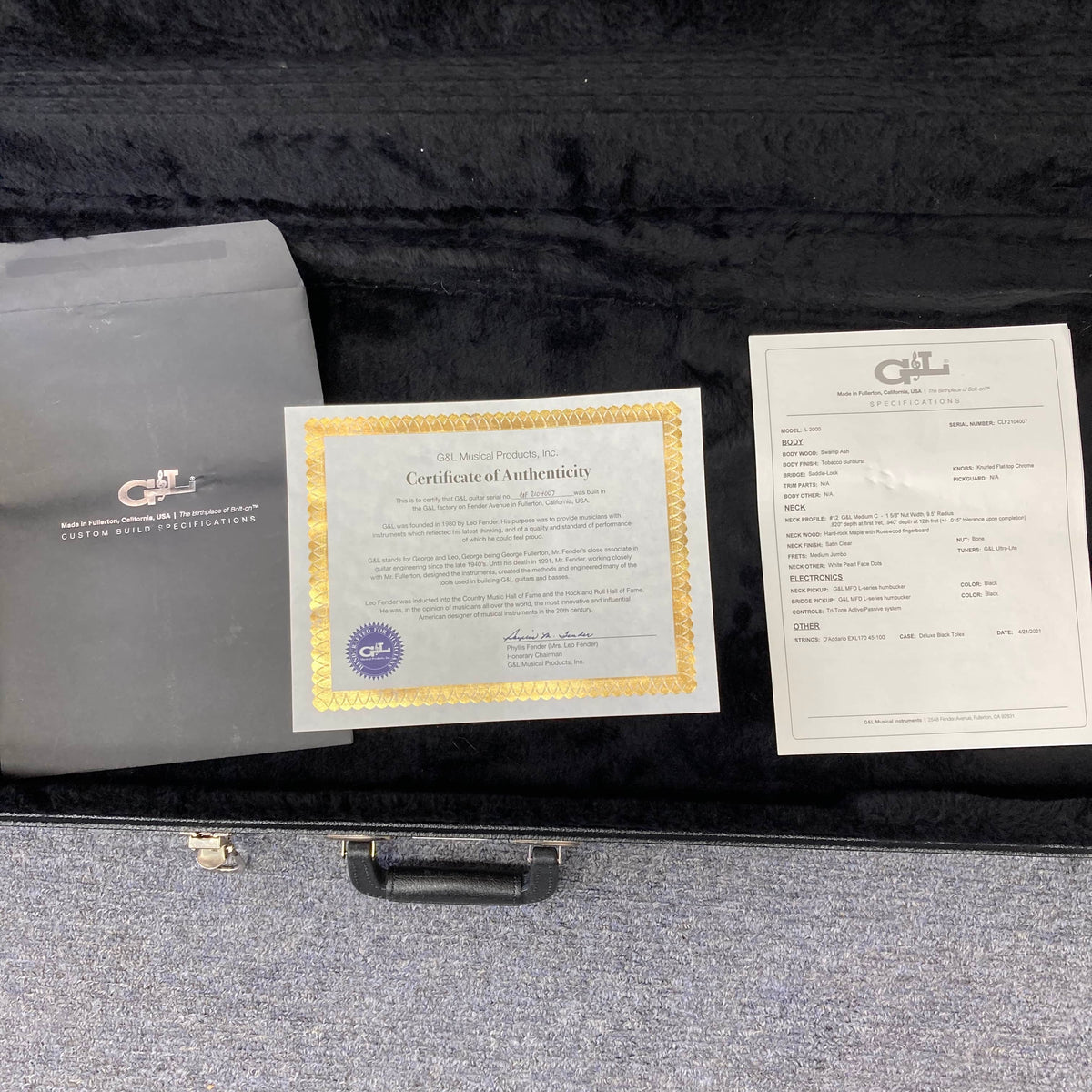 A certificate of authenticity, spec sheet, and black envelope are shown inside an open G&L L-2000 USA Tobacco Sunburst Bass w/Case Used Consign on a gray carpet, highlighting its classic finish.