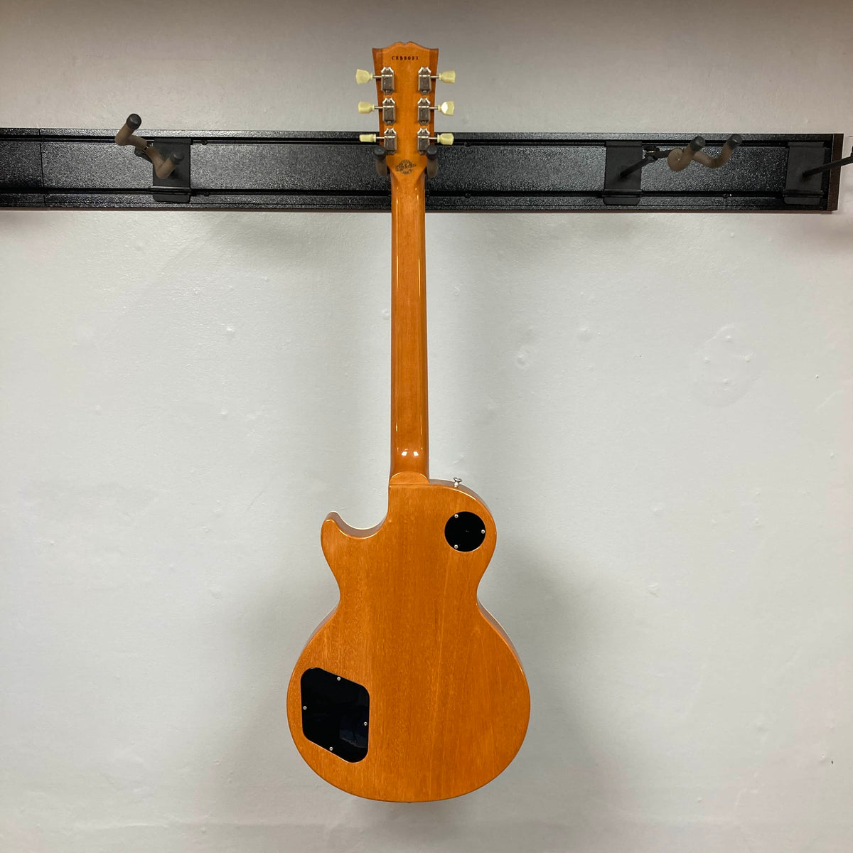 A Gibson Custom Shop Class 5 Les Paul electric guitar with a natural wood finish hangs on a wall mount, seen from the back with its control cavity covers removed and tuning pegs visible.