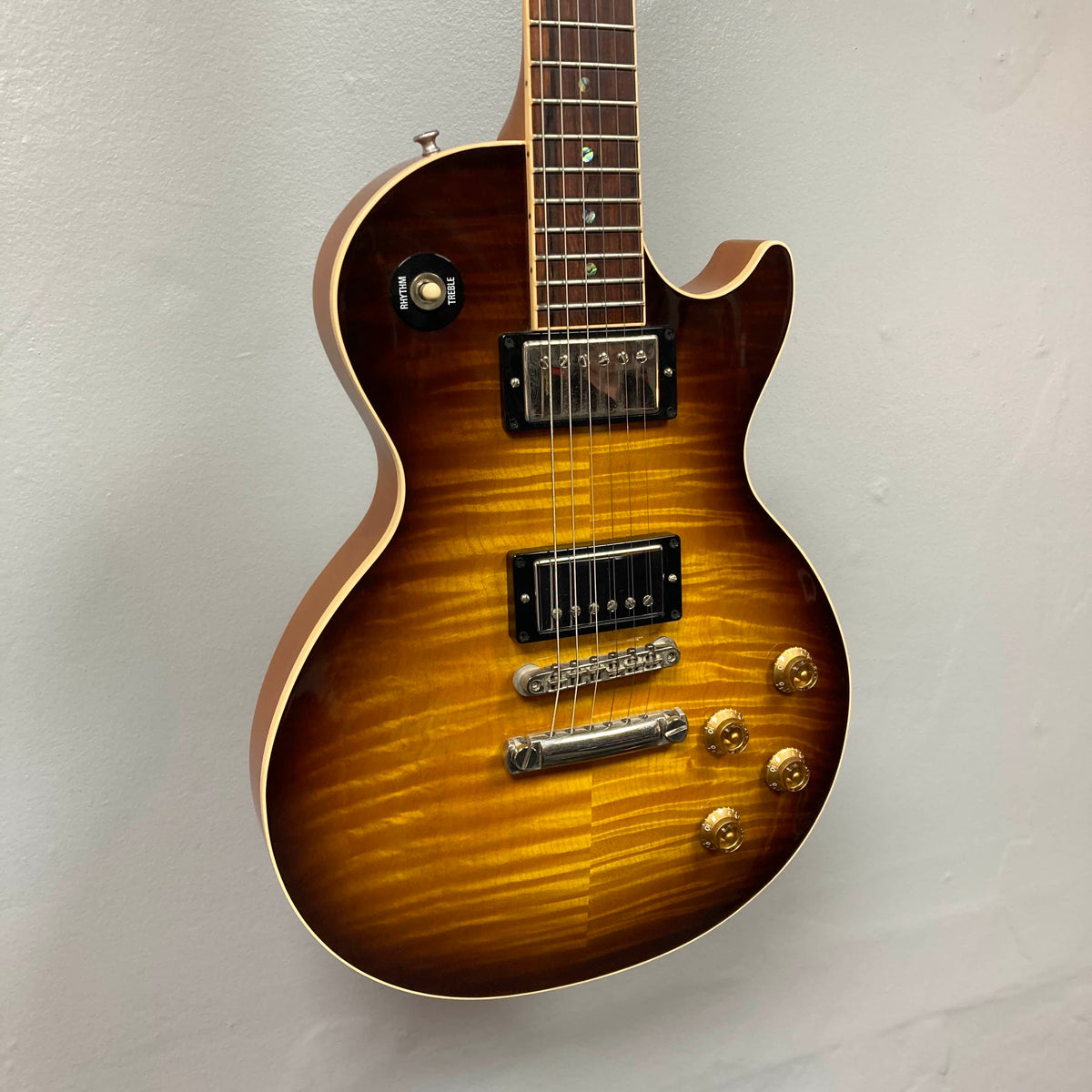 A Gibson Custom Shop Class 5 Les Paul electric guitar with a flame maple top, dual humbuckers, four gold control knobs, and a toggle switch is displayed against a plain light-colored wall.