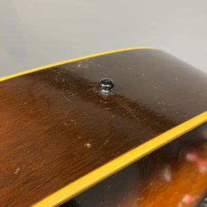Close-up of a Gibson J-45 1957 Serial U-8664-1 acoustic guitar body by Gibson, showing its brown wood finish with scratches, black strap button, yellow binding, and classic slope shoulder design.
