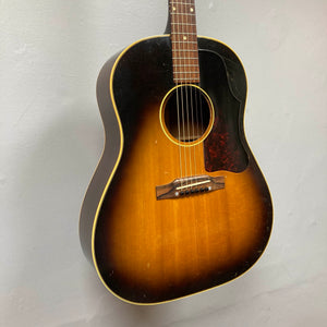 A Gibson J-45 1957 acoustic guitar with a classic sunburst finish and slope shoulder design hangs on a light-colored wall.
