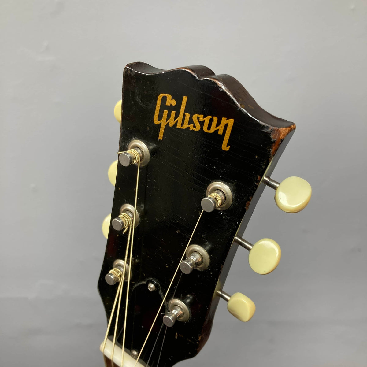 Close-up of the Gibson J-45 1957 Serial U-8664-1 headstock in black, featuring six tuning pegs and a gold Gibson logo, highlighting the classic design of this iconic vintage guitar against a plain gray background.