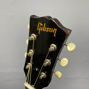 Close-up of the Gibson J-45 1957 Serial U-8664-1 headstock in black, featuring six tuning pegs and a gold Gibson logo, highlighting the classic design of this iconic vintage guitar against a plain gray background.