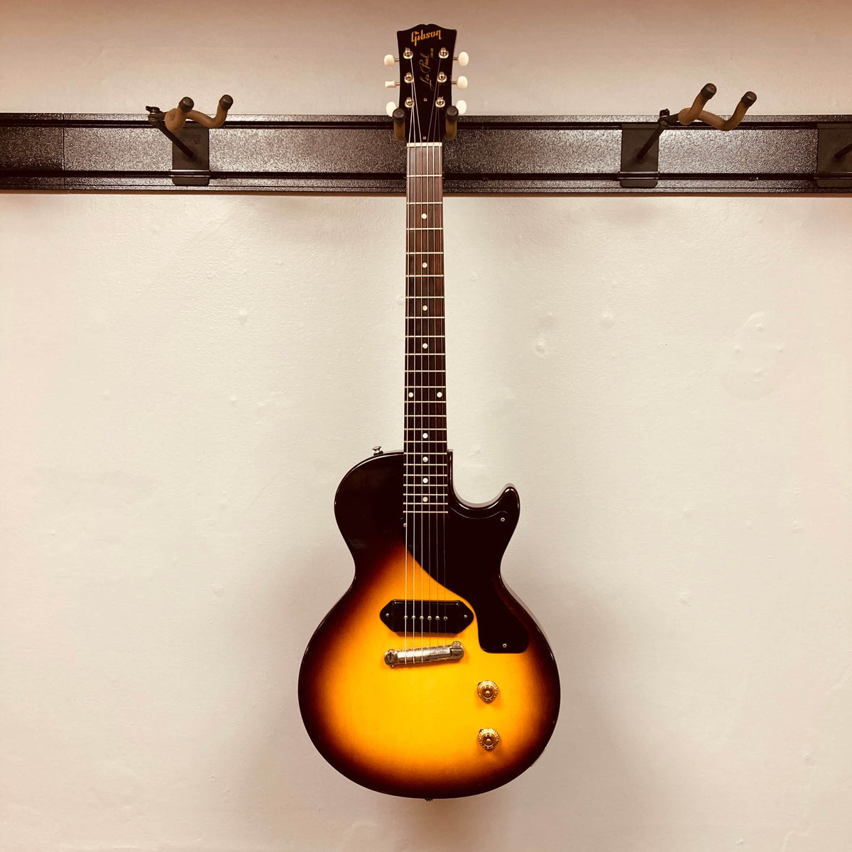 A Gibson Les Paul Junior 1957 Sunburst electric guitar with a single cutaway and P-90 pickup by Gibson hangs on a wall-mounted rack against a plain white background.
