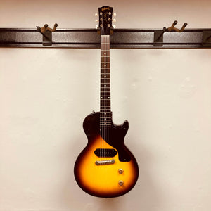 A Gibson Les Paul Junior 1957 Sunburst electric guitar with a single cutaway and P-90 pickup by Gibson hangs on a wall-mounted rack against a plain white background.