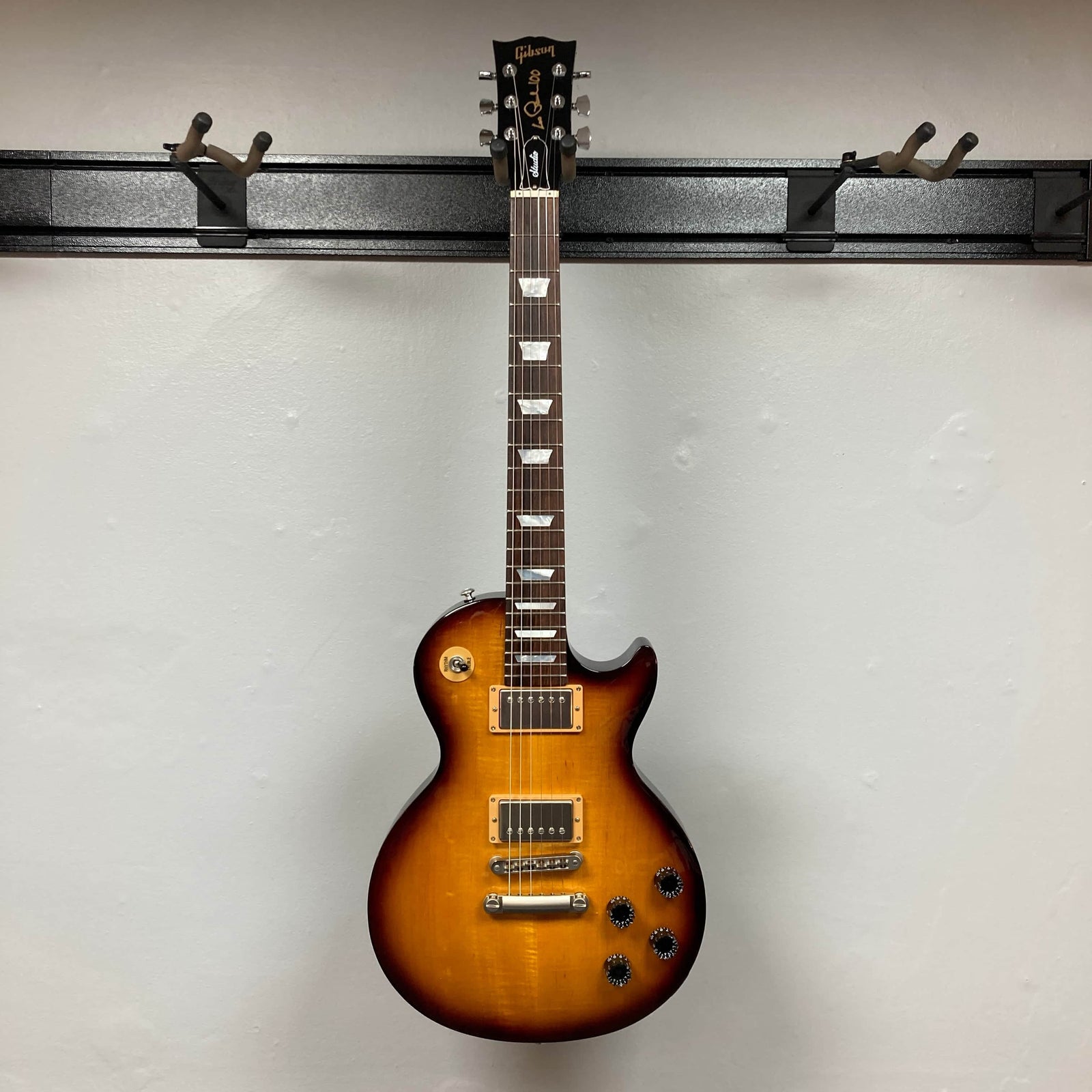 A Gibson Les Paul Studio 2015 Desert Burst electric guitar with two humbucker pickups, four knobs, locking tuners, and a single cutaway, shown against a plain light gray wall. Includes case. Brand: Gibson. A Gibson Les Paul Studio 2015 Desert Burst electric guitar with two humbucker pickups, four knobs, locking tuners, and a single cutaway, shown against a plain light gray wall. Includes case. Brand: Gibson.
