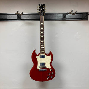 A Gibson SG Standard Heritage Cherry yr 2000 Consign electric guitar by Gibson, featuring a white pickguard, hangs on a wall-mounted rack against a plain white wall, highlighting its USA-made craftsmanship.