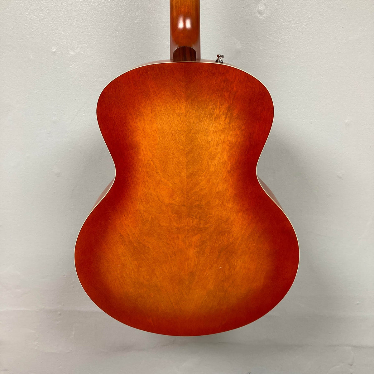 The back of the Godin 5th Avenue Jumbo HB Memphis Sun guitar, with its glossy sunburst orange wood finish, gleams against a plain light-colored wall.