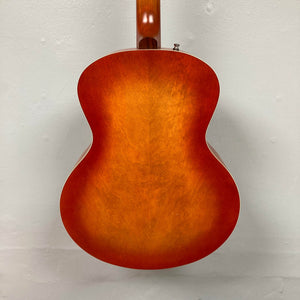 The back of the Godin 5th Avenue Jumbo HB Memphis Sun guitar, with its glossy sunburst orange wood finish, gleams against a plain light-colored wall.