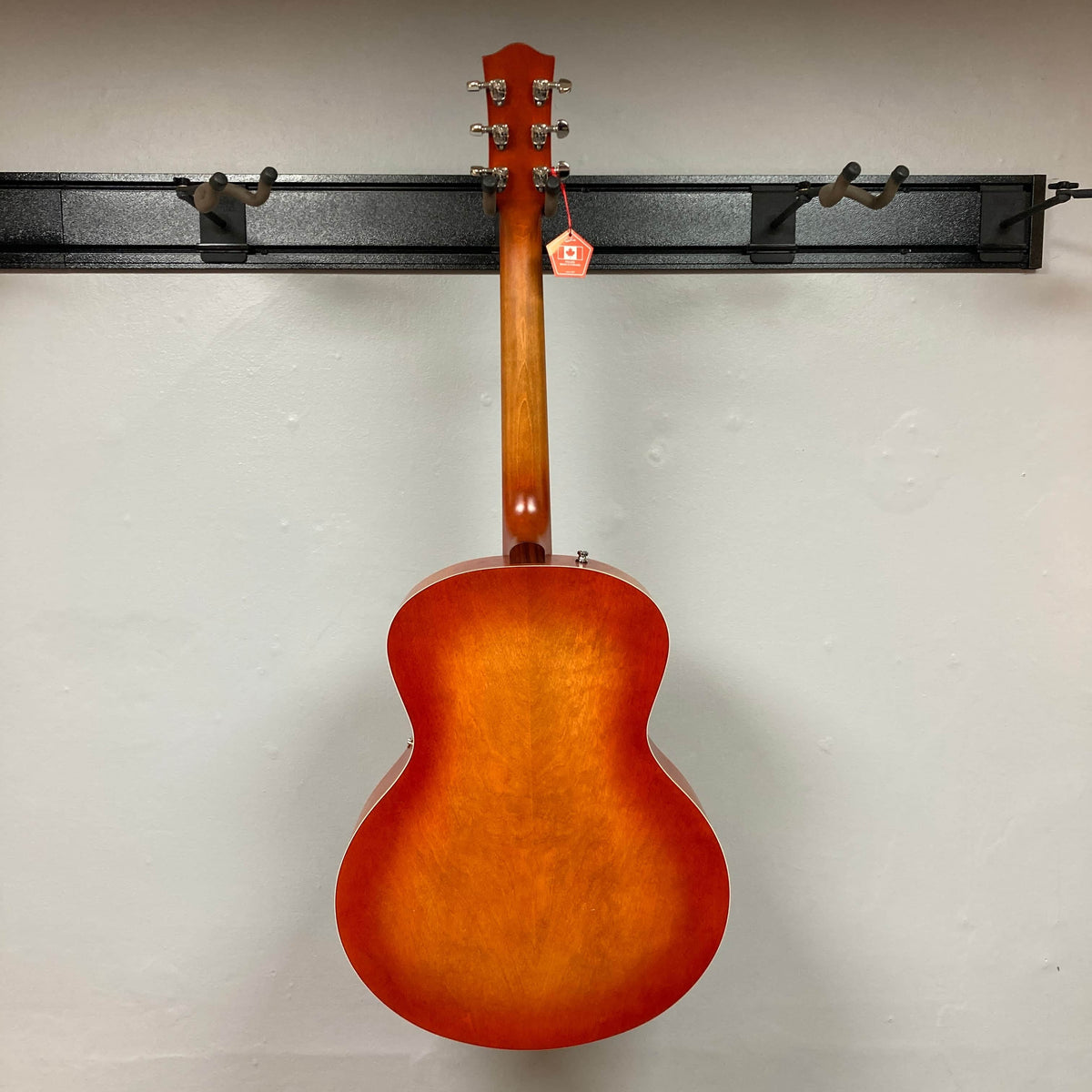 A Godin 5th Avenue Jumbo HB Memphis Sun guitar by Godin hangs on a wall-mounted rack, viewed from the back with a red tag attached to a tuning peg.