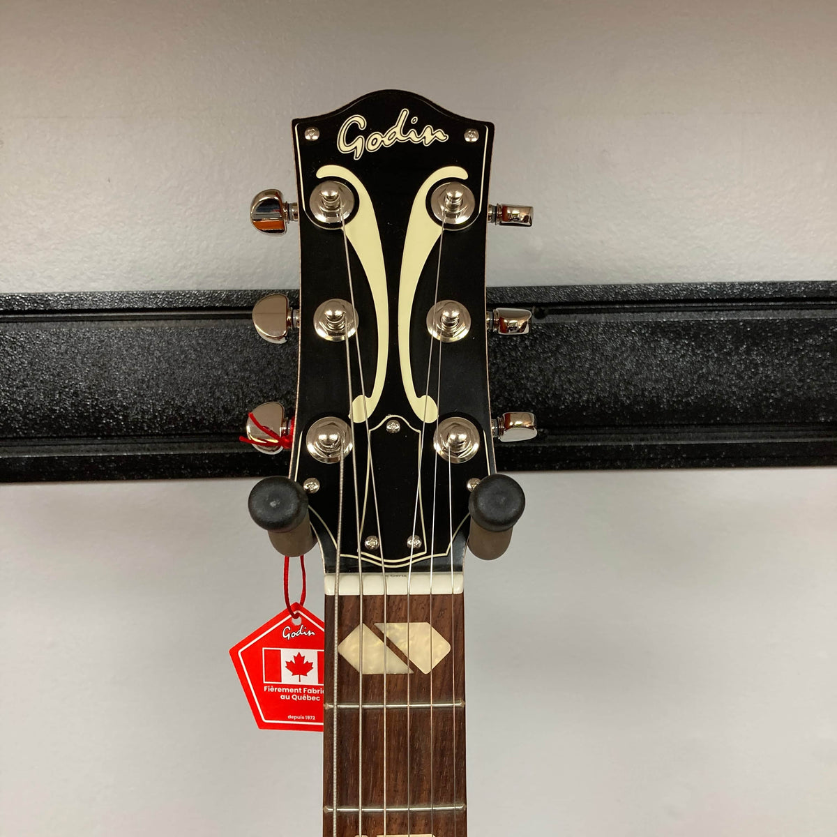 The Godin 5th Avenue Jumbo HB Memphis Sun guitar's headstock shows tuning pegs, a decorative inlay, and a red "Made in Canada" tag while mounted on a wall hanger.