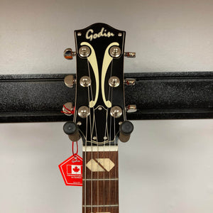 The Godin 5th Avenue Jumbo HB Memphis Sun guitar's headstock shows tuning pegs, a decorative inlay, and a red "Made in Canada" tag while mounted on a wall hanger.