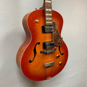 A Godin 5th Avenue Jumbo HB Memphis Sun guitar with a sunburst finish, two humbuckers, Seymour Duncan pickup, two knobs, pickguard, and f-holes hangs against a plain wall.