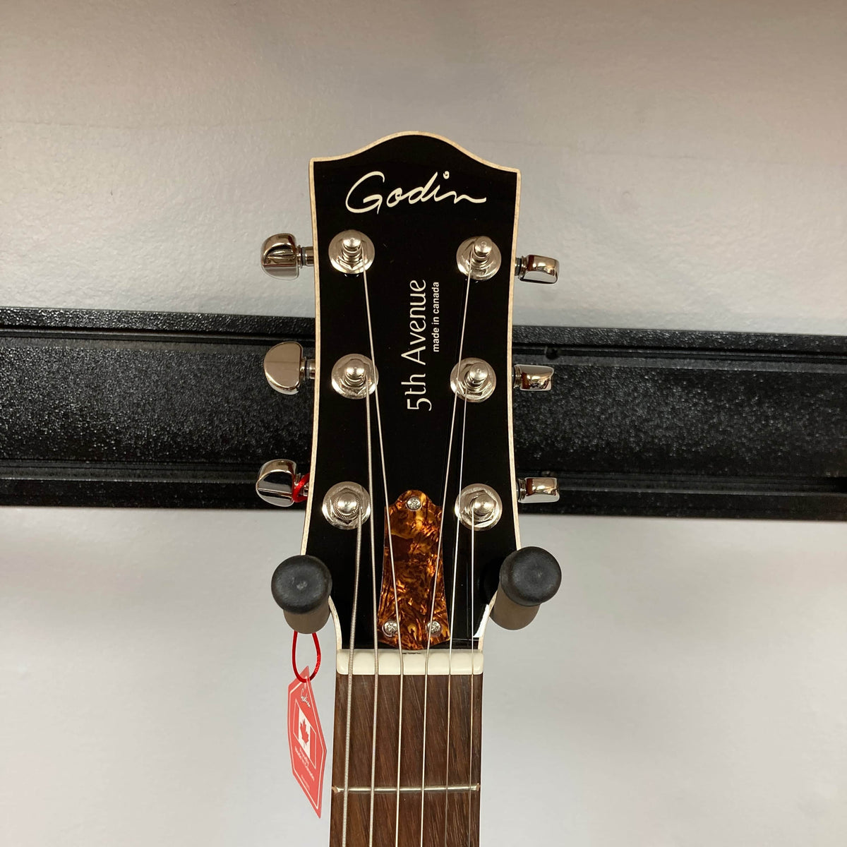 Close-up of a Godin 5th Avenue Kingpin P90 Cognac Burst SF headstock, displaying tuning pegs, Godin logo, model name, and a red tag on one peg. The guitar is shown mounted on a black wall hanger.