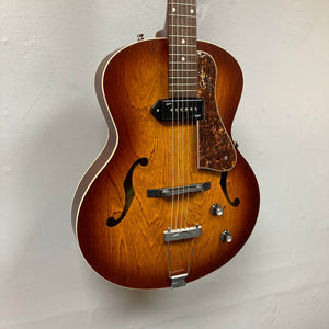 A Godin 5th Avenue Kingpin P90 Cognac Burst SF archtop guitar with two f-holes, a single P90 pickup, vintage tone, tortoiseshell pickguard, and metallic tailpiece hangs against a light-colored wall.