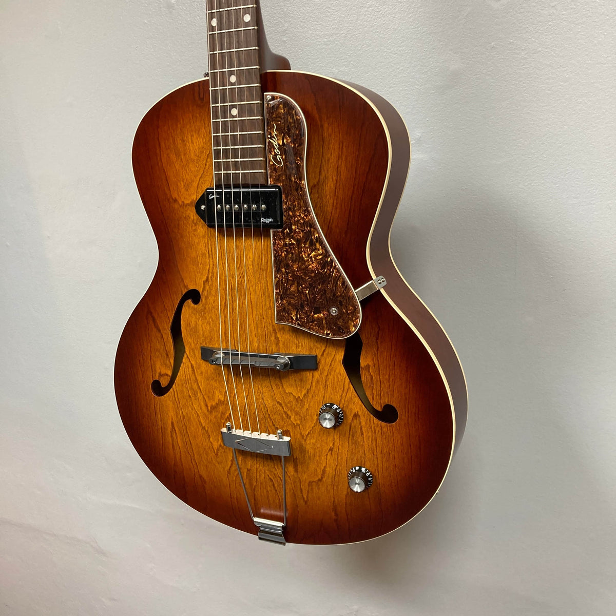 A Godin 5th Avenue Kingpin P90 Cognac Burst SF archtop electric guitar with a single pickup, f-holes, and tortoiseshell pickguard hangs on a light wall, ready to deliver its classic Godin sound.