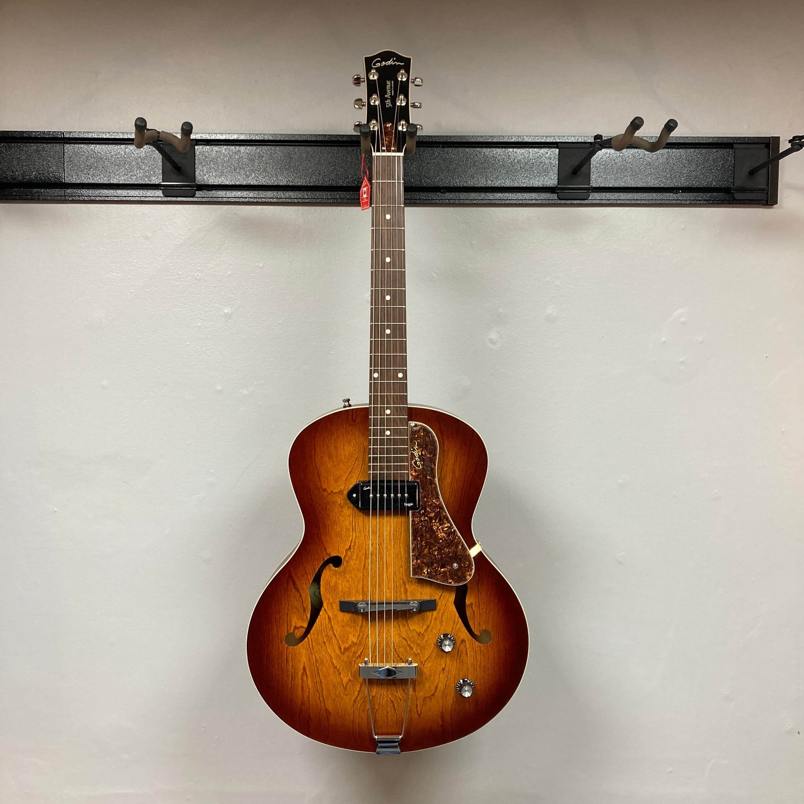 A Godin 5th Avenue Kingpin P90 Cognac Burst SF archtop electric guitar with dual f-holes, two knobs, a wooden pickguard, and visible wood grain hangs against a light wall, ready to deliver classic vintage tones. A Godin 5th Avenue Kingpin P90 Cognac Burst SF archtop electric guitar with dual f-holes, two knobs, a wooden pickguard, and visible wood grain hangs against a light wall, ready to deliver classic vintage tones.
