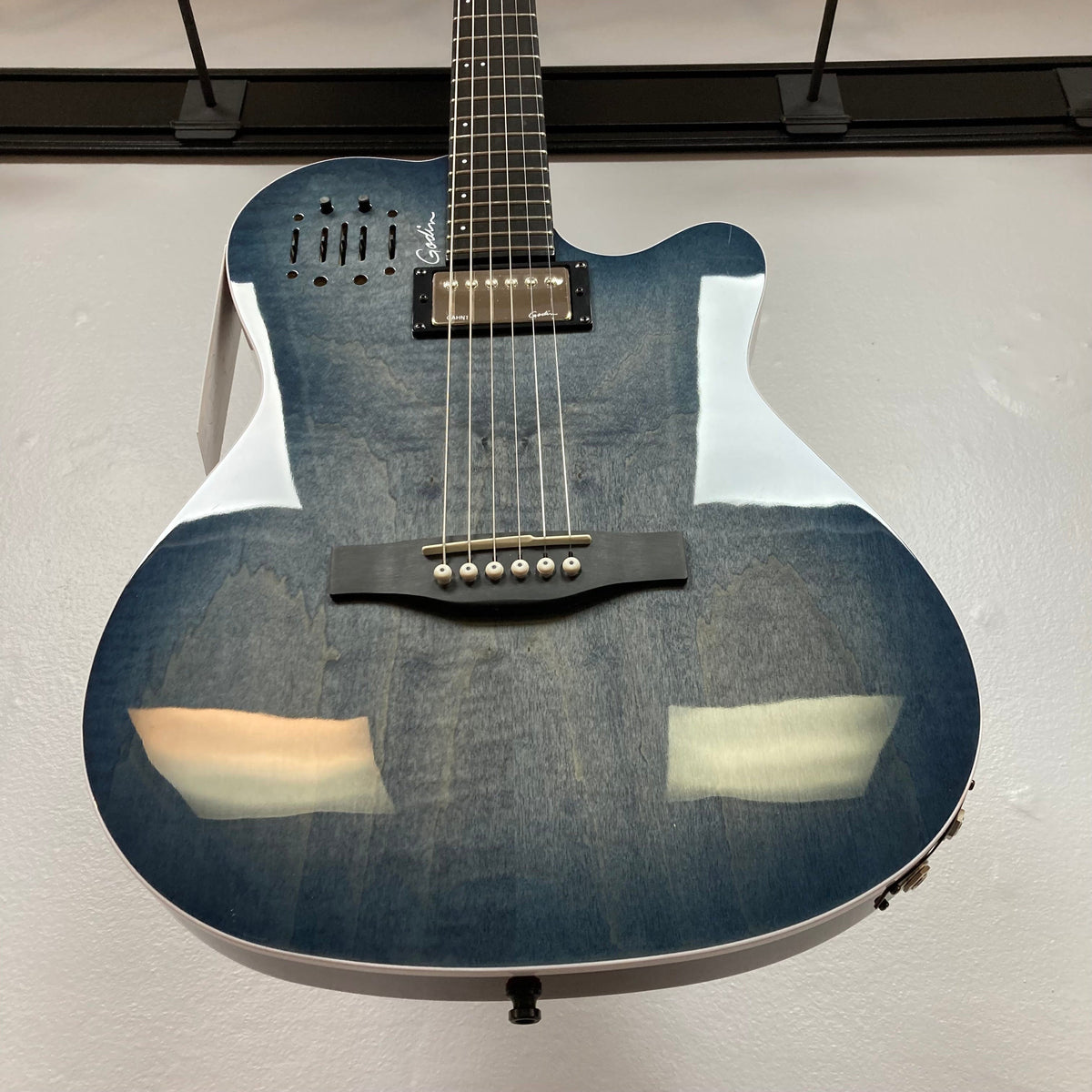 A close-up of the Godin A6 Ultra Denim Blue Flame SF with Bag, showing its cutaway body, six strings, black bridge, and glossy finish as it hangs vertically on a wall.