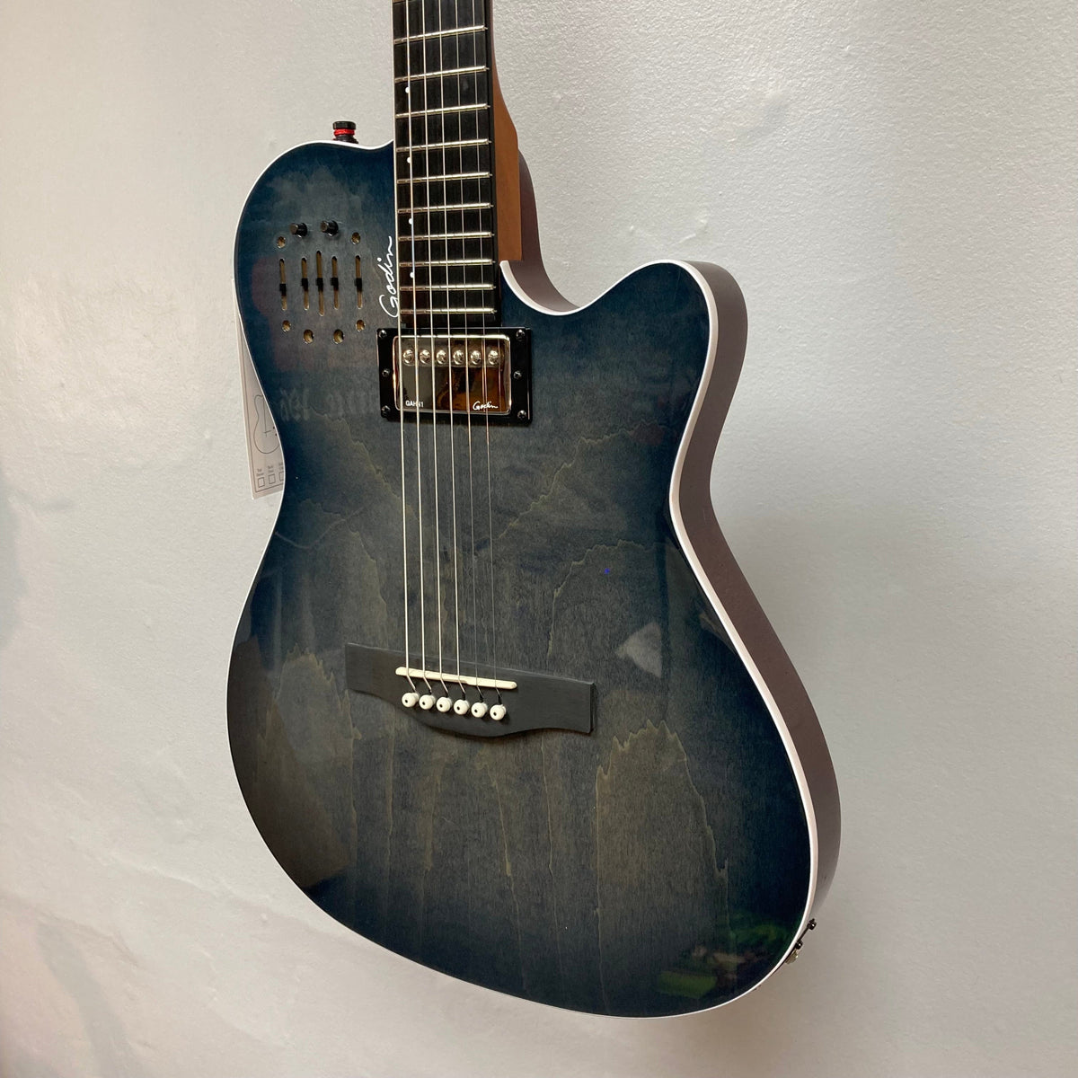 A close-up of the Godin A6 Ultra Denim Blue Flame SF acoustic electric guitar, featuring a cutaway body, six strings, single pickup, and bridge, mounted against a plain white wall. Brand: Godin. Includes bag.