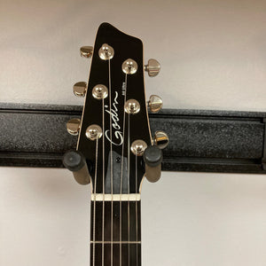 A close-up of the Godin A6 Ultra Denim Blue Flame SF with Bag headstock displays six tuning pegs, the Godin logo, and part of a wall-mounted holder against a light-colored wall.