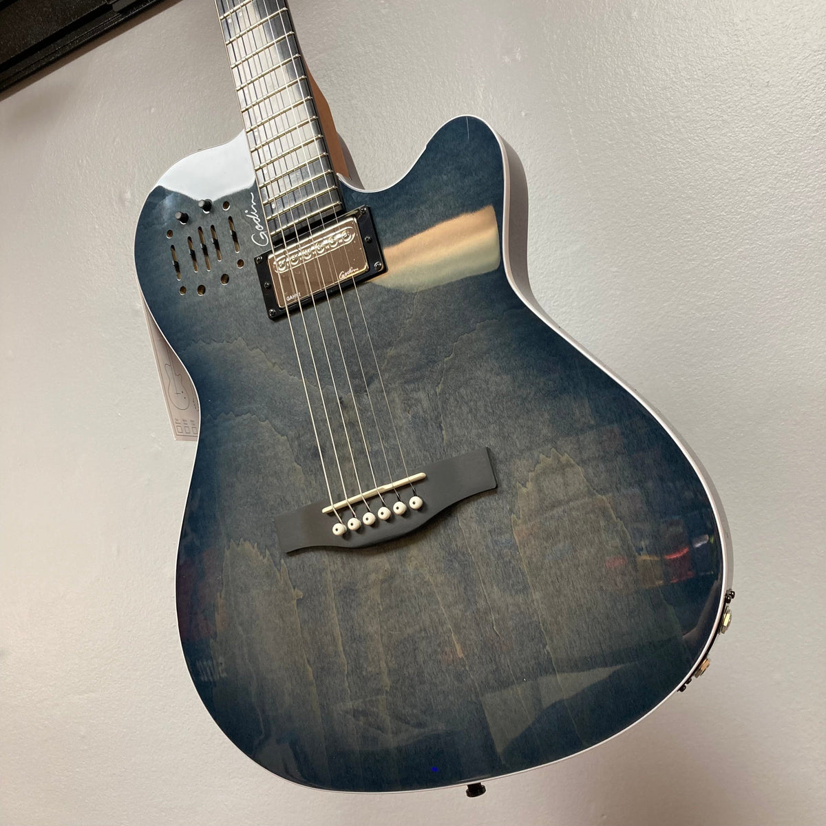 A Godin A6 Ultra Denim Blue Flame SF acoustic electric guitar with a glossy, wood-grain finish hangs on the wall, showing its body, bridge, pickup, and neck. Includes gig bag.