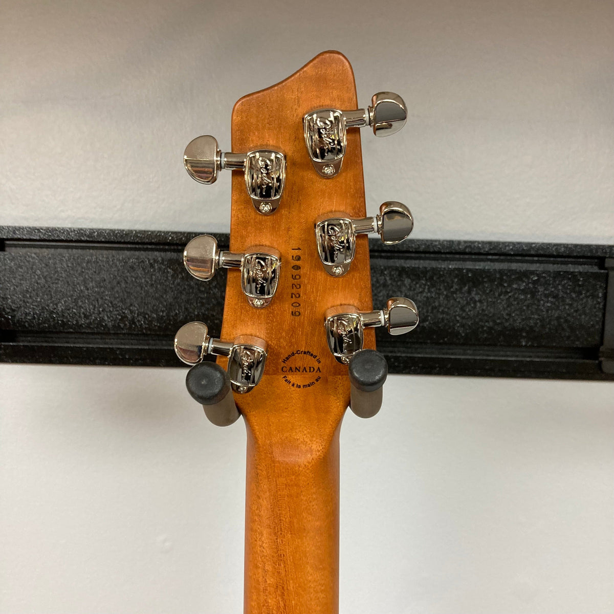 The back of the Godin A6 Ultra Denim Blue Flame SF with Bag shows six tuning pegs, serial number, and a "Made in Canada" stamp. The guitar by Godin rests on a wall mount with a black padded yoke.
