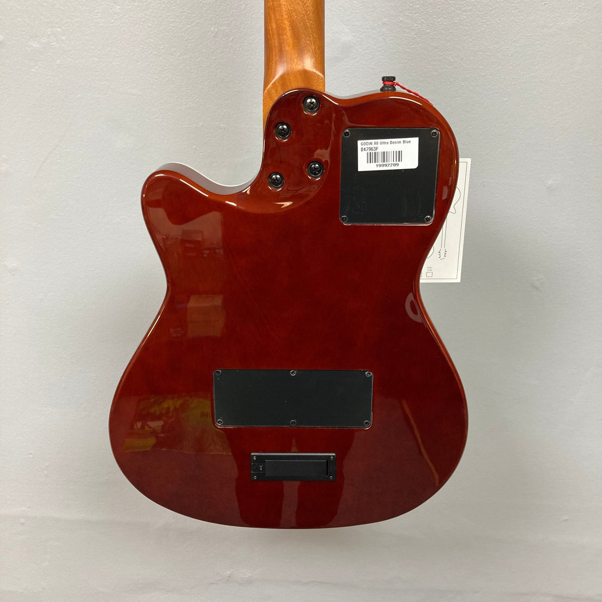 The Godin A6 Ultra Denim Blue Flame SF with Bag features a glossy Denim Blue Flame finish, two black back plates, control knobs, barcode sticker, and a wood neck. Shown from the back against a plain light wall. Brand: Godin.
