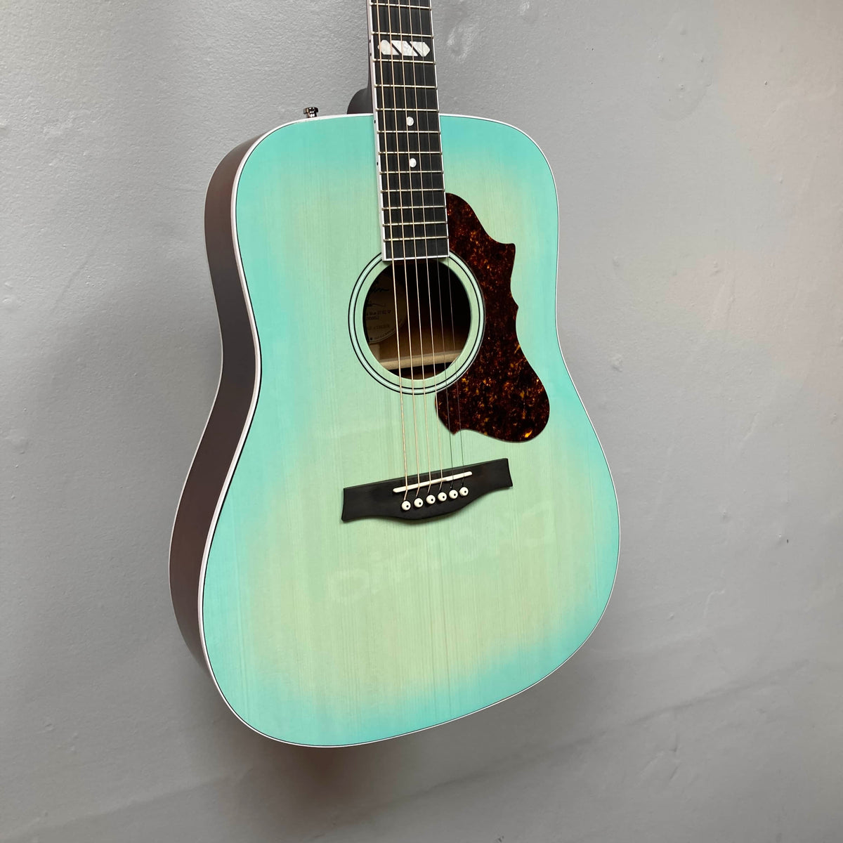 A Godin Imperial Laguna Blue GT EQ SF acoustic guitar with gigbag is hanging on a light gray wall.