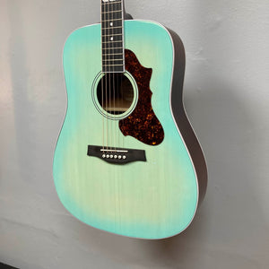 A Godin Imperial Laguna Blue GT EQ SF acoustic guitar with a tortoiseshell pickguard and LR Baggs Anthem system hangs against a plain gray wall.