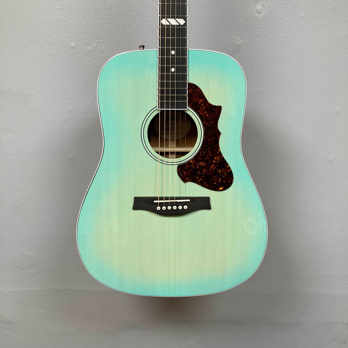A Godin Imperial Laguna Blue GT EQ SF acoustic guitar with gigbag hangs against a plain light gray wall.