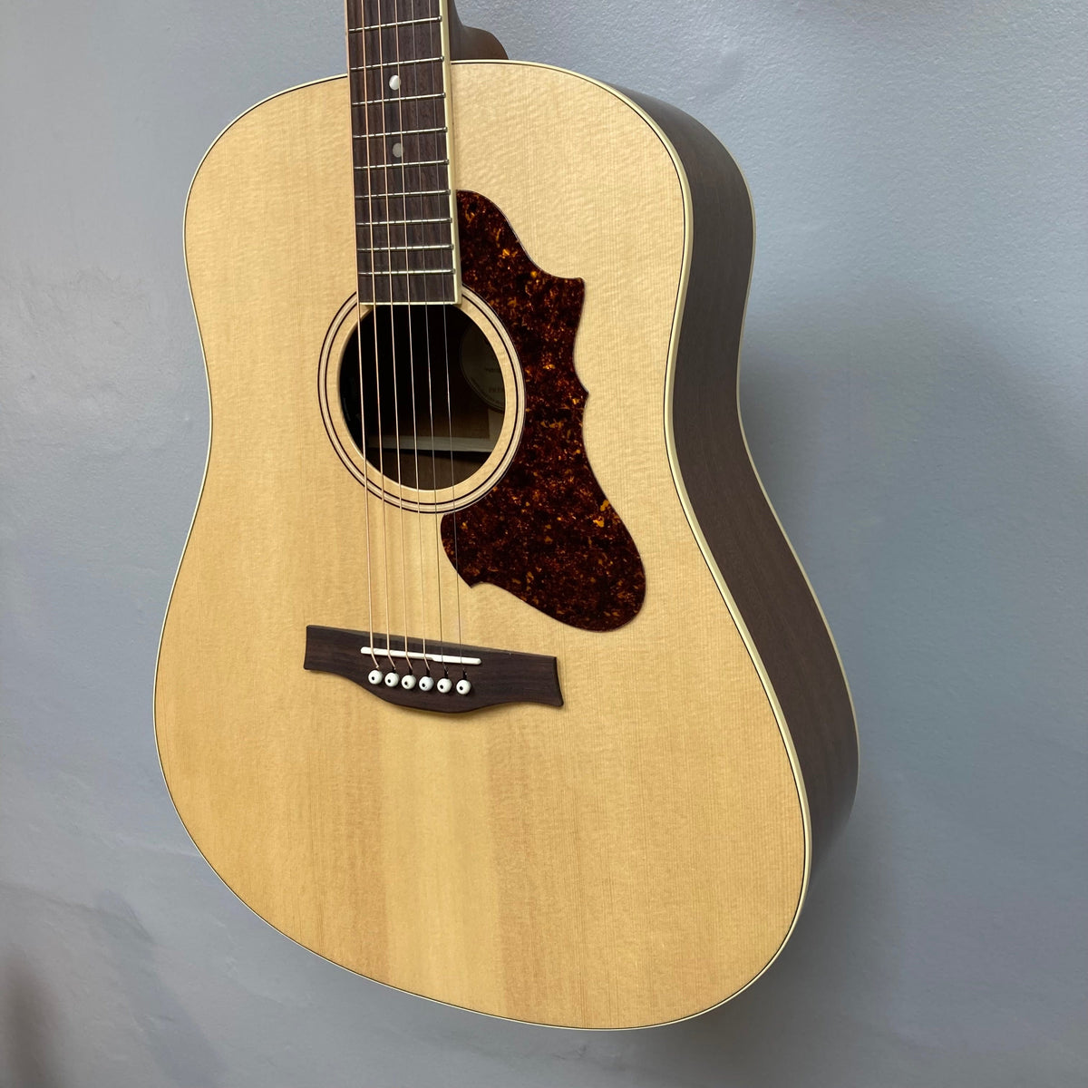Godin Metropolis Natural RN GT EQ acoustic guitar with mahogany back, solid spruce gloss top, and rosewood fretboard, displayed on a wall.