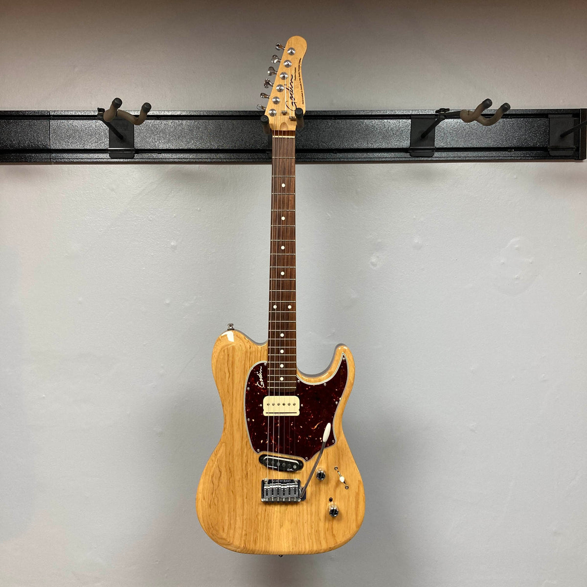 The Godin Passion Custom Swamp Ash Electric Guitar w/ GigBag Consign, by Godin, features a natural wood finish, dark red pickguard, and six tuning pegs. It hangs on a wall-mounted guitar rack against a light gray wall and is equipped with Seymour Duncan P-Rail pickups for exceptional tonal versatility.