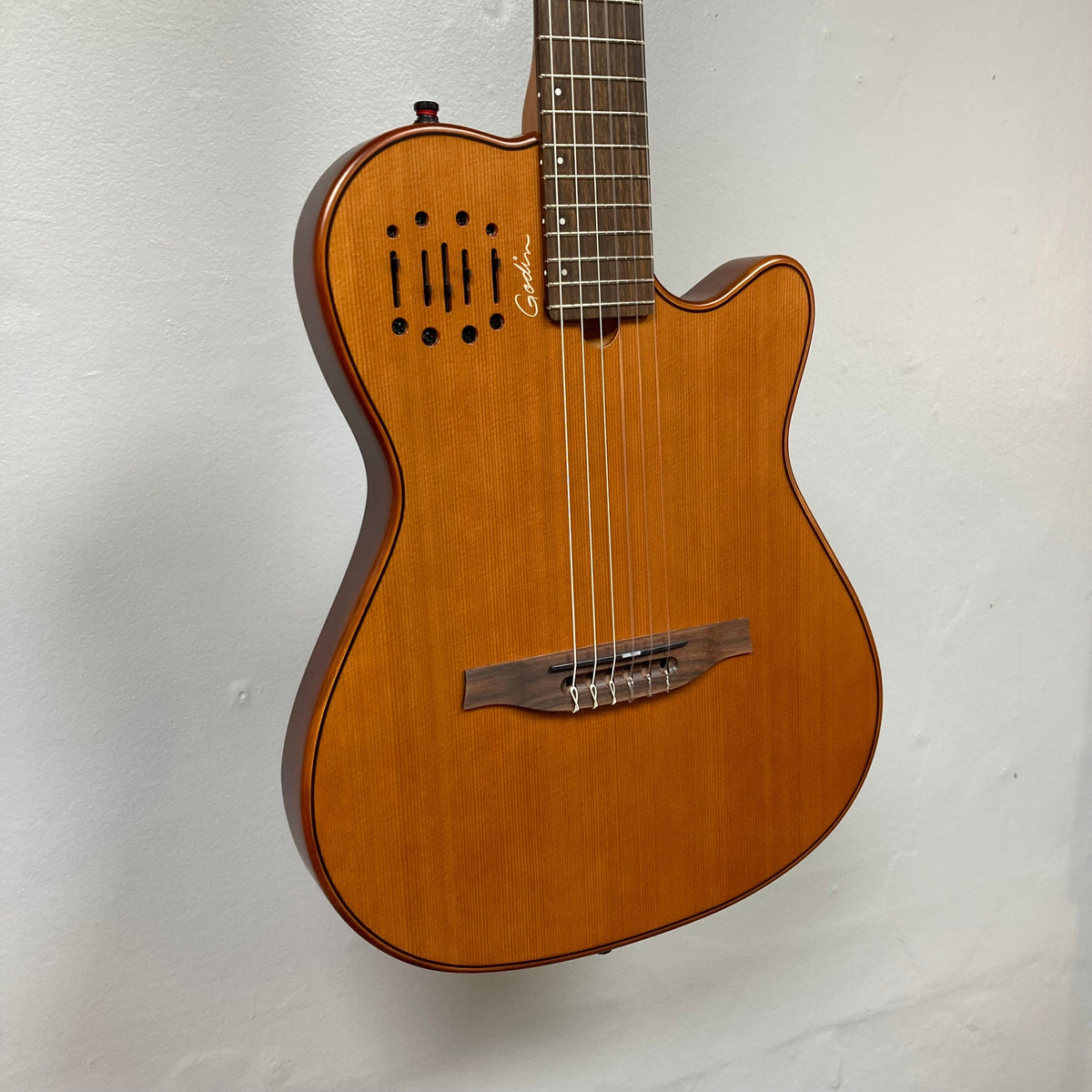 Godin Prototype Ltd Mundial Dark Cedar nylon-string guitar with visible strings and neck, displayed against a white wall.