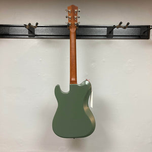 A Godin Radium Matte Green electric guitar hangs on a wall-mounted rack, displayed with its back facing outward against a plain white wall.