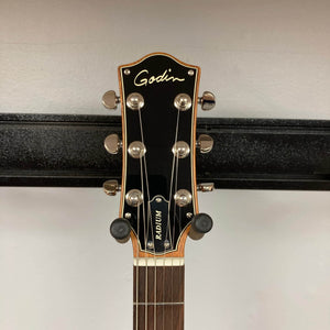 Close-up of the headstock of the Godin Radium Matte Green guitar, featuring tuning pegs, the Godin logo, and part of the neck against a plain wall background.