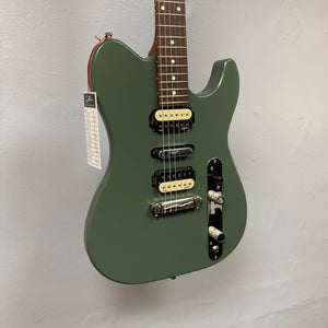 A Godin Radium Matte Green electric guitar with Godin Zebra Humbuckers (H-S-H pickup configuration), tag on the headstock, hangs against a light gray wall.