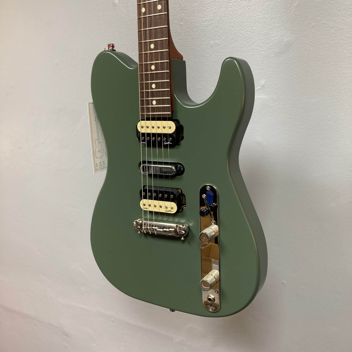 A Godin Radium Matte Green electric guitar with Zebra Humbuckers, tune-o-matic bridge, and chrome knobs hangs against a white wall.