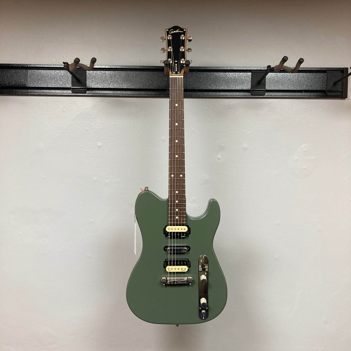 A Godin Radium Matte Green electric guitar with an H-S-H pickup setup and Godin Zebra Humbuckers hangs on a wall-mounted rack against a plain white background.
