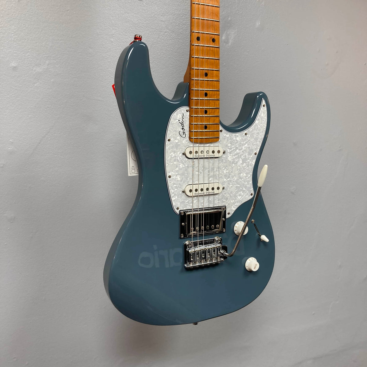 A Godin Session T-Pro electric guitar with a maple neck, white pearloid pickguard, H-S-S pickups, and a tremolo bridge hangs against a plain gray wall. Comes with a branded bag.