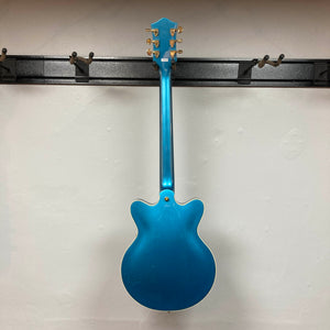 A Gretsch G2655TG Streamliner Riviera Blue Refurb semi-hollow electric guitar is seen from the back as it hangs on a wall-mounted rack with empty hooks against a plain white wall.
