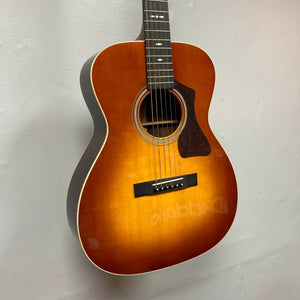 A Guild Gad-30reasb acoustic guitar with a sunburst finish, crafted from solid Indian rosewood, is hanging on a white wall, displaying its front body and fretboard.