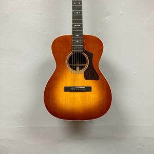 The Guild Gad-30reasb w/Case, an orchestra-size acoustic-electric guitar with a sunburst finish, is hanging on a plain, light-colored wall and features a Fishman Matrix pickup for exceptional amplified sound.