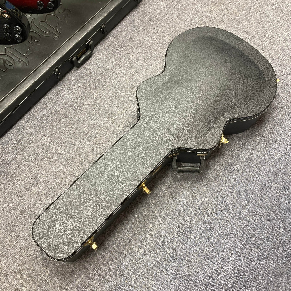 A black Guild USA F-512 w/OHSC 2005 Circa guitar case with gold latches lies on a gray carpeted floor.