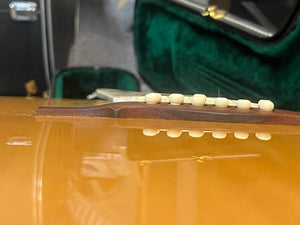 Close-up side view of a Guild USA F-512 w/OHSC 2005 Circa jumbo 12-string acoustic guitar bridge lifting from the body, with bridge pins and green velvet case lining visible. Crafted by Guild in Tacoma, Washington.