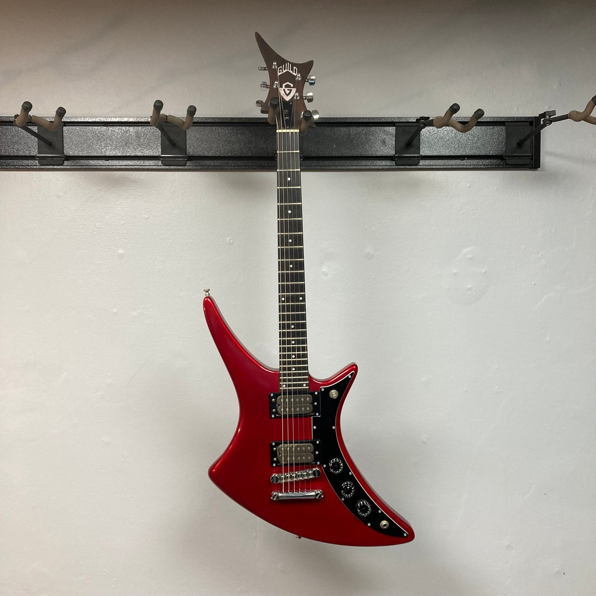 Guild USA X-79 Candy Apple Red electric guitar on a hook, featuring a unique body shape and 24-fret fingerboard, with a case included.