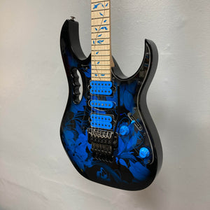 The Ibanez JEM77P Steve Vai Signature from Guitars on Main features a black and blue floral design, blue pickups and knobs, and a maple fretboard with blue inlays, displayed against a white wall.