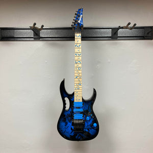 The Ibanez JEM77P Steve Vai Signature from Guitars on Main features a blue floral finish, maple neck with distinctive inlays, and matching blue headstock, displayed on a wall-mounted holder.