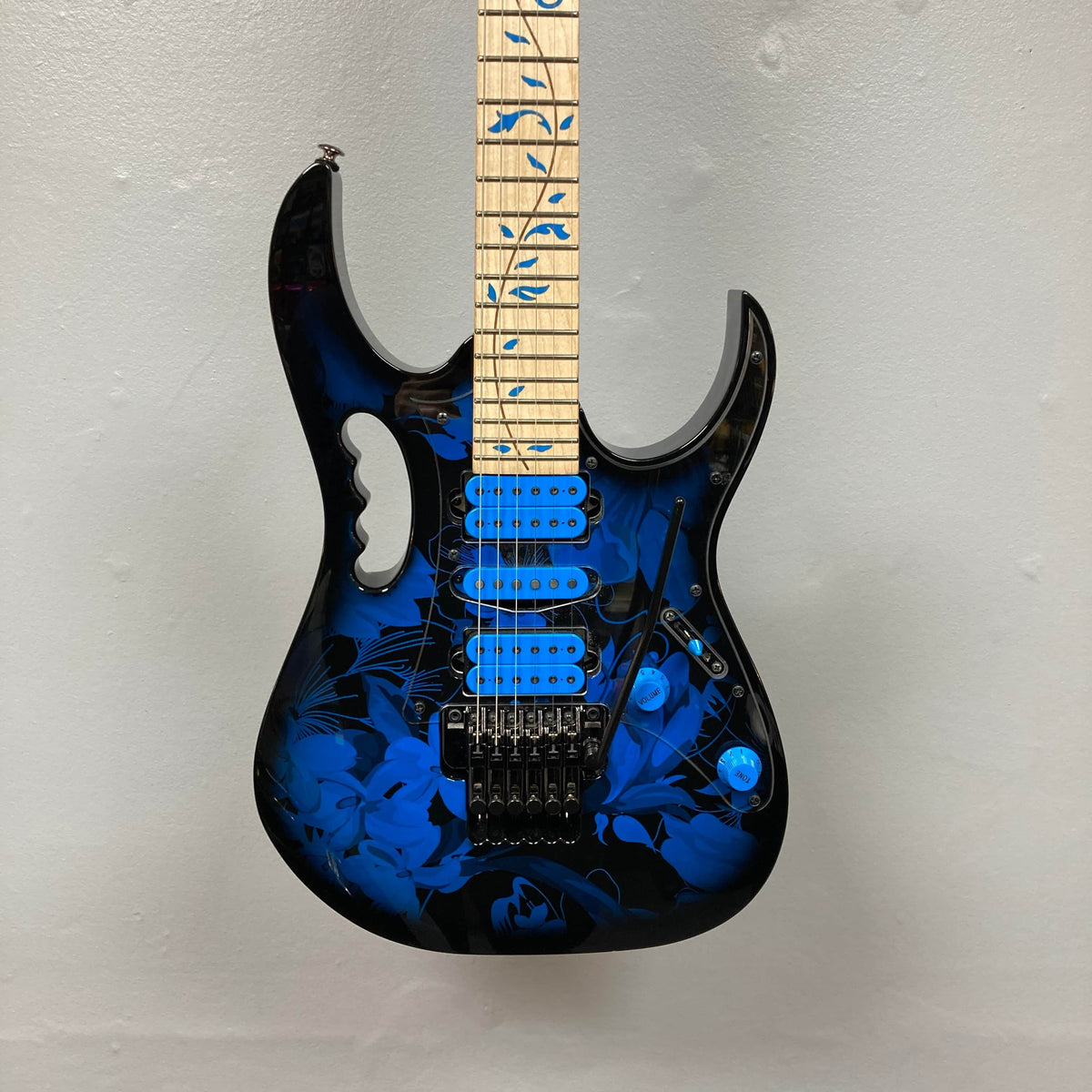 The Ibanez JEM77P Steve Vai Signature from Guitars on Main features a black body with blue floral patterns, blue pickups, and a maple neck with blue inlays, displayed against a light gray wall.